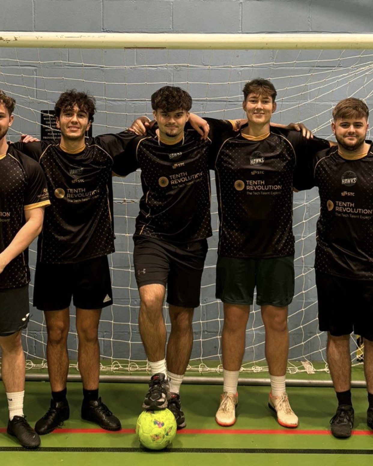 A five-a-side football team in matching black and gold kit, standing in front of the goal after a match